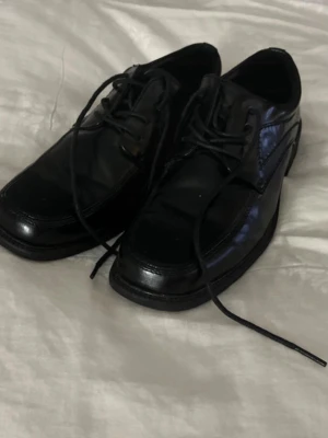 Svarta klassiska snörskor - Säljer ett par svarta snörskor med fyrkantig tå och robust sula. Skorna har en stilren design och är tillverkade i blankt material, troligen syntet eller läderimitation. Passar perfekt till dig som vill ha en clean och tidlös look.