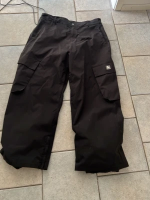 Svarta täckbyxor från Harlaut - Säljer ett par svarta täckbyxor från Harlaut med bred passform och stora cargofickor på sidorna. Perfekta för vinterns alla äventyr. Byxorna har klassisk look och praktiska detaljer som gör dem både snygga och funktionella. Använda 1 Säsong, mycket bra skick, det enda som är är lite slitet är längst ner på byxan då det är långa så man råkar gå på det