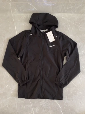 Svart Nike vindjacka med huva - Snygg svart vindjacka från Nike med reflekterande detaljer och klassisk logga på bröstet. Jackan har huva, hel dragkedja framtill och elastiska muddar vid ärmsluten. Tillverkad i lätt och tåligt material, perfekt för blåsiga dagar.