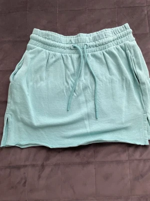 Ljusgrön kortkjol med snörning - Snygg ljusgrön kortkjol med snören i midjan. Kjolen har två fickor och små slitsar i sidorna för en avslappnad look. Perfekt för varma dagar och enkel att matcha med olika toppar.