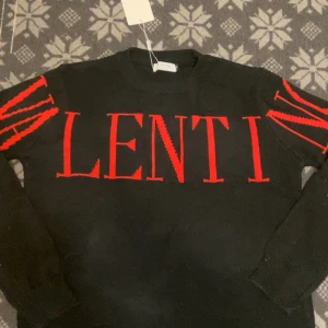 Svart stickad tröja från Valentino - Säljer en svart stickad tröja från Valentino med stora röda bokstäver över bröstet och röda detaljer på ärmarna. Tröjan har rund halsringning och långa ärmar. Perfekt för dig som gillar statement-plagg och exklusiva märken.