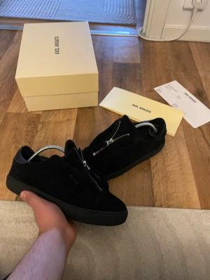 Svarta sneakers från Axel Arigato - Snygga svarta sneakers från Axel Arigato i mocka med diskret logga på sidan och dragkedja på ovansidan. Skorna har en chunky svart sula och klassisk låg profil. Perfekta för dig som gillar stilrena och moderna detaljer.