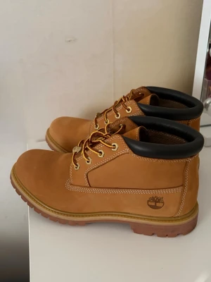 Timberland klassiska kängor  - Säljer ett par klassiska Timberland boots i ljusbrunt nubuckläder med svart vadderad kant och robust gummisula. Skorna har guldfärgade öljetter och gulbruna snören. Perfekta för dig som gillar streetwear och tidlös stil.
