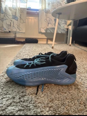 Adidas blå sneakers med meshdetaljer - Säljer ett par snygga Adidas sneakers i svart och ljusblått. Skorna har en futuristisk design med meshdetaljer på sidan, blå snören och Adidas-logga på hälen. Ovandelen är i textil och syntet, sulan är i gummi. Perfekta för dig som gillar streetwear och unik stil.