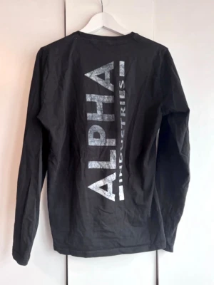Svart långärmad topp Alpha Industries - Svart tröja från Alpha Industries. Stort tryck på ryggen och liten logga fram. Storlek S. Använd men fin. Inget ytludd, ej blekt, trycket fint - "sliten look".  Den är i fint skick.