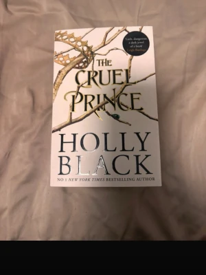 The Cruel Prince av Holly Black - The Cruel Prince är en fantasybok på engelska av Holly Black. Omslaget är vitt med guldfärgade och svarta detaljer, inklusive en krona och grenar. Boken är i pocketformat och har tydliga titeldetaljer i guld och svart. Den är på engelska!!!
