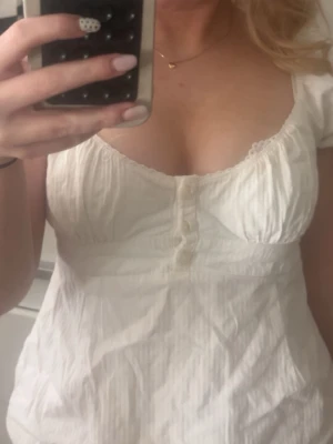 Vit blus med knappar och puffärm - Söt vit blus med korta puffärmar och v-ringning. Toppen har dekorativa knappar framtill och lätt rynkade detaljer vid bysten. Tillverkad i ett tunt, strukturerat bomullstyg som ger en luftig känsla. Perfekt för en fräsch och trendig look.