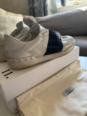 Valentino vita och mörkblå sneakers - Säljer ett par snygga sneakers från Valentino med vit läderbas och bred mörkblå läderdetalj över mitten. Skorna har klassisk låg profil, platta snören och nitar på sulan. Perfekt för dig som gillar exklusiva märken och stilren design. En liten fläck se bild 4 .