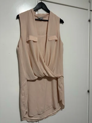 Beige omlott ärmlös linnetopp - Säljer en snygg beige ärmlös linnetopp med omlottdesign och dekorativa fickdetaljer framtill. Toppen har en avslappnad passform och är tillverkad i ett tunt, mjukt material som passar perfekt till sommaren.