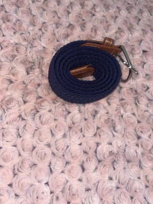 Blått flätat bälte  - Snyggt marinblått flätat bälte med detaljer i brunt skinn och silverfärgat spänne. Perfekt accessoar för att lyfta din outfit med en klassisk touch. Bältet har vita sömmar på skinnpartierna och är flexibelt tack vare det flätade materialet.