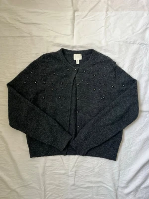 Mörkgrå kofta med pärlor från H&M - Mysig mörkgrå kofta från H&M med rund halsringning och dekorativa pärlor på framsidan. Koftan är stickad och har långa ärmar samt knappar framtill. Perfekt för dig som vill ha något extra till din outfit. Diskuterar gärna pris!✨