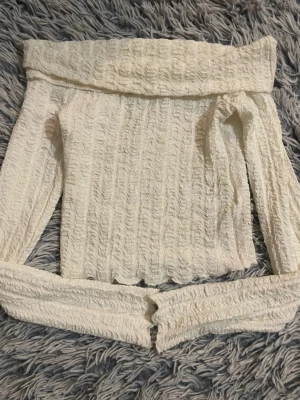 Vit offshoulder topp med struktur - Trendig vit offshoulder topp med lång ärm och snyggt strukturerat tyg. Toppen har en croppad passform och bred kant upptill som ger en cool look. Perfekt att styla med höga jeans eller kjol för en modern vibe.