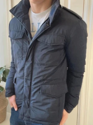 Mörkblå vindjacka med fickor - Snygg mörkblå vindjacka med hög krage och fyra stora fickor framtill. Jackan har axelklaffar och stängs med dragkedja och knappar. Materialet är lätt och vindtätt, perfekt för blåsiga dagar. Passar dig som gillar en stilren och funktionell look.