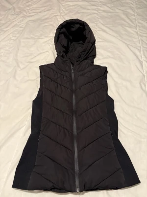 Svart dunväst med huva - Snygg svart dunväst med dragkedja framtill och vadderad design i chevronmönster. Västen har en stor huva och sidopaneler i stretchigt material för extra komfort. Perfekt för lager-på-lager och streetwear-stil. Är i perfekt skick och andvänd 1 gång! Storlek S
