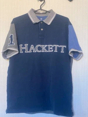 Blå och grå Hackett t-shirt - Snygg t-shirt från Hackett i marinblått och grått med kontrasterande ärmar och stor logga över bröstet. Klassisk pikékrage med knappar och nummerdetalj på ena ärmen. Perfekt för en sportig och avslappnad stil.