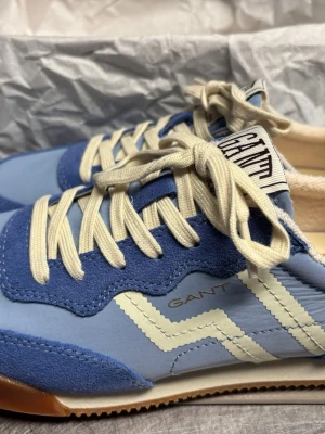 Blå och vita sneakers från Gant - Snygga sneakers från Gant i ljusblått och mörkblått med vita detaljer och krämfärgade snören. Skorna har en retroinspirerad design med vågig sula och Gant-logga på sidan och plösen. Materialmix av mocka och textil för en cool look.