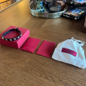 Valentino svart nitar armband - Snyggt svart armband från Valentino med silverfärgade pyramidformade nitar. Tillverkat i läder och har ett justerbart spänne. Kommer med original ask och dustbag. Perfekt accessoar för en edgy och trendig stil.