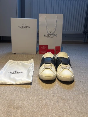Valentino Garavani Open sneakers vit/blå - Snygga Valentino Garavani Open sneakers i vitt skinn med bred marinblå rem över snörningen. Klassisk rund tå och platt sula med mönstrad undersida. Guldlogga på plösen och detaljer i vitt och blått. Kommer med originalkartong och dustbag.