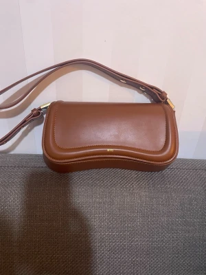 Brun axelväska från ATP Atelier - Säljer en snygg brun axelväska från ATP Atelier i slätt läder. Väskan har en unik vågformad kant, justerbar axelrem och guldfärgade detaljer. Perfekt för dig som gillar stilrena accessoarer med en twist.