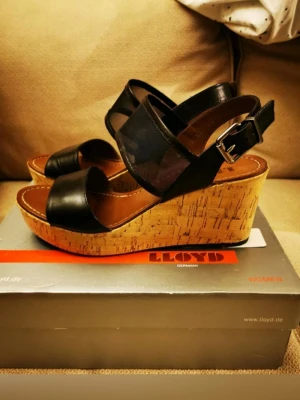 Svarta kilklackspumps från Lloyd - Snygga svarta pumps från Lloyd med kilklack i kork och breda remmar i läder och mesh. Justerbart spänne vid ankeln och öppen tå ger en modern look. Perfekta för dig som vill ha både höjd och komfort i din stil. Helt nya och oanvända i box med tags