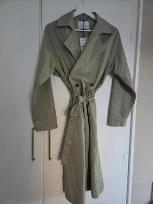 Grön trenchcoat från Stradivarius - Grön trenchcoat från Stradivarius med krage, knytskärp i midjan och dubbelknäppning framtill. Helt ny, aldrig använd, prislapp kvar.