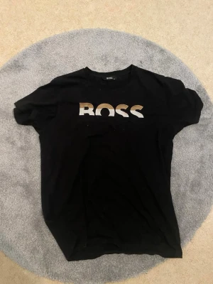 Svart BOSS t-shirt med logotyp - Säljer en svart t-shirt från BOSS med stort logotryck i vitt, silver och guld på bröstet. T-shirten har rund hals och korta ärmar, perfekt för dig som vill ha en stilren och trendig look. Materialet känns mjukt och bekvämt mot huden.
