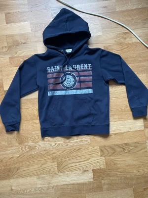 Mörkblå hoodie från Saint Laurent - Snygg mörkblå hoodie från Saint Laurent med tryck framtill i vitt och rött. Klassisk känguruficka och justerbar huva med snören. Tillverkad i mjukt bomullsmaterial för en bekväm känsla. Perfekt för dig som vill ha en stilren och trendig look.