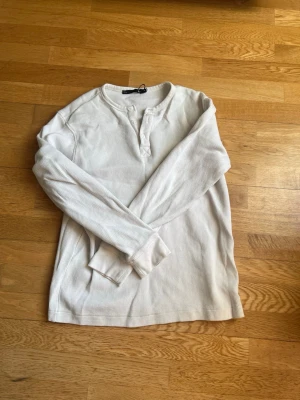 Vit långärmad henley från 157 - Klassisk vit långärmad henleytröja från 157 med ribbad struktur och knappar vid halsen. Tillverkad i mjukt och bekvämt material, perfekt för lager på lager eller som den är. Enkel och stilren design som passar till det mesta.