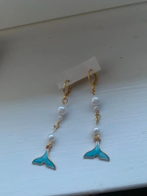 Guldörhängen med pärlor och blå fenor - Säljer ett par unika örhängen med guldfärgade detaljer, vita pärlor och blå emaljerade fenor längst ner. Örhängena har en hängande design och ger en cool havsinspirerad vibe. Perfekta för dig som gillar statement-smycken och vill sticka ut.