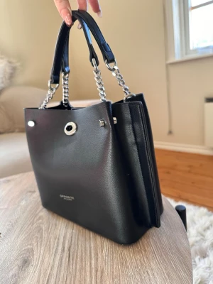 Svart handväska från Opposite - Stilren svart handväska från Opposite med kedjedetaljer på handtagen och silverfärgade metalldetaljer. Väskan är tillverkad i slätt konstläder och har en boxig form med två handtag. Perfekt för dig som vill ha en snygg och praktisk väska.