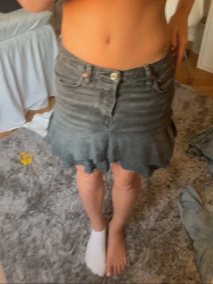 Grå jeanskjol med volangkant - Säljer en grå jeanskjol med volangkant och råa kanter. Kjolen har klassiska fem fickor, knapp och dragkedja framtill. Perfekt för dig som gillar en edgy och trendig stil.