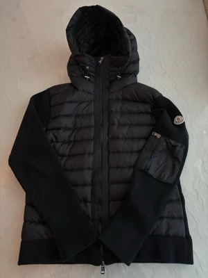 Svart Moncler cardiganjacka med luva - Snygg svart cardiganjacka från Moncler med quiltad front och stickade ärmar. Jackan har luva med dragsko, dragkedja framtill och en ficka med Moncler-logga på ärmen. Perfekt för dig som vill ha både stil och komfort. OBS: Kan diskutera om priset vid snabba affärer. 
