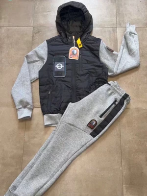 Grått loungewear-set med svart väst - Snyggt loungewear-set från PGS med grå mjukisbyxor och matchande huvtröja med svart vadderad väst. Setet har dragkedjor på både väst och byxor samt ribbade muddar. Perfekt för chill dagar hemma eller när du vill vara bekväm.