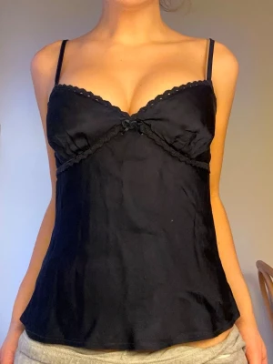 Marinblå spetsdetaljerad top från Subdued💙 - Snygg marinblå top från Subdued med tunna axelband och spetskant vid bysten. Materialet är mjukt och glansigt, troligen satin eller polyester. Dekorativ rosett framtill och lätt utsvängd nederdel. Perfekt för dig som gillar stilrena underkläder med en feminin touch.💙