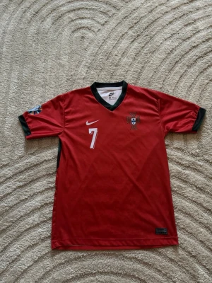 Portugal fotbollströja Ronaldo #7 Nike - Röd Portugal fotbollströja från Nike med nummer 7 och Ronaldo på ryggen. Tröjan har svarta detaljer vid krage och ärmslut, samt landslagets emblem på bröstet. Perfekt för dig som älskar fotboll och vill stötta Portugal.