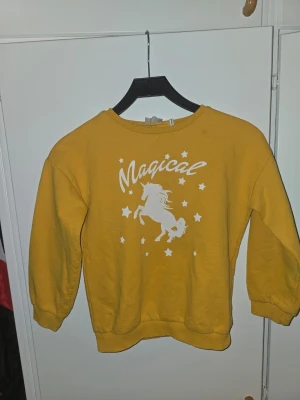 Gul sweatshirt med enhörning - Cool gul sweatshirt med tryck av en vit enhörning, texten 'Magical' och små stjärnor på bröstet. Tröjan har rund hals, ribbade muddar vid ärmar och nederkant. Perfekt för dig som gillar magiska motiv och vill sticka ut lite extra.