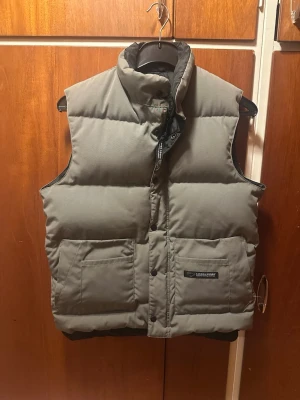 Olivgrön väst från Canada goose - Snygg olivgrön dunväst från Canada, den har både dragkedja och knappar. Perfekt för lager-på-lager och håller dig varm under kyliga dagar. Färgen visas bäst på bilden där den är på. Jag är 180cm och runt 70kg. Hör av vid några frågor.