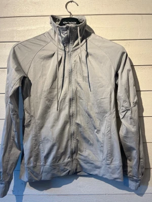 Ljusgrå träningsjacka från Casall - Ljusgrå träningsjacka från Casall med hel dragkedja, hög krage och dragsko. Jackan har långa ärmar, figurnära passform och diskret logga på bröstet. Perfekt för träning eller som lager på väg till gymmet.