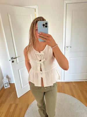 Vit ärmlös blus med rosetter - Söt vit blus i bomull med volangkant och tre dekorativa rosetter framtill. Modellen är ärmlös och har en lätt utsvängd passform som ger en luftig känsla. Perfekt för varma dagar och enkel att styla med olika byxor eller kjolar.