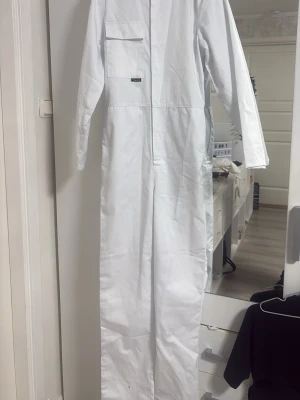 Studentoveroll  - Vit långärmad overoll med klassisk krage, bröstficka och helknäppning framtill. Tillverkad i ett tåligt bomullsliknande material och har resår i ryggen för bättre passform. Den är i en väldigt vit färg så den ser inte gul eller så ut utan den är väldigt vit