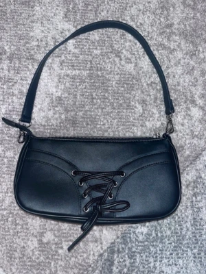 Svart axelväska från Even&Odd - Snygg svart axelväska från Even&Odd i fejkläder med cool snörningsdetalj framtill. Väskan har en enkel, avlång form och dragkedja upptill. Perfekt accessoar för att lyfta din outfit med en edgy touch.