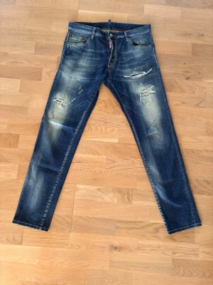Blå slitna jeans från Dsquared2 - Säljer ett par blå jeans från Dsquared2 med snygga slitningar och distressed detaljer. Jeansen har smal passform, klassisk femficksdesign och coola slitna partier på låren och knäna. Perfekta för en trendig och avslappnad look.99% bomull 
