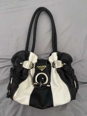 Svartvit handväska Prada  - Snygg svartvit handväska från Prada (kan ej bekräfta äkthet) med två flätade handtag och detaljer i silverfärgad metall. Väskan har ett coolt mönster med breda ränder, dekorativa spännen och kedja framtill. Tillverkad i läder med tydlig Prada-logga på framsidan. Defekter förekommer, går nog att måla över dem