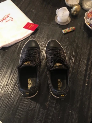 Svarta Fendi sneakers med monogram - Snygga svarta sneakers från Fendi med klassiskt FF-monogram i brunt och svart. Skorna har svarta snören, rund tå och detaljer i läder. Fendi-logga syns på plösen och innersulan. Perfekta för dig som gillar lyxiga streetwear-vibbar.