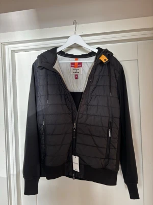 Svart pufferjacka/cardigan från Parajumper - Snygg svart pufferjacka från Parajumpers med huva och dragkedja framtill. Jackan har quiltad front och ryggparti, mjuka ärmar och muddar samt en gul detalj vid halsen. En sportig, modern look.
