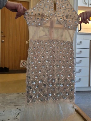 Glittrig beige kortklänning med paljetter - Säljer en glittrig beige kortklänning med transparenta partier, täckt av stora paljetter och pärlor i ett rutmönster. Klänningen har hög hals, är ärmlös och avslutas med fjädrar längst ner för extra wow-effekt. Perfekt för dig som vill sticka ut!