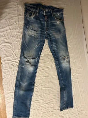Blå slitna skinny jeans Dsquared2 - Säljer ett par blå skinny jeans från Dsquared2 med slitningar, hål på knäna och coola detaljer som färgstänk och patch baktill. Jeansen har klassisk femficksmodell och är tillverkade i denim med stretch för skön passform.