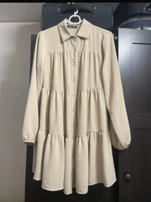 Beige Boohoo Volangklänning  - Luftig beige kortklänning från Boohoo med volanger och knappar framtill. Klänningen har långärmade och flowiga ärmar med resår vid handlederna och klassisk krage. Perfekt för en avslappnad och trendig look.