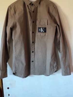  Calvin Klein overshirt skjorta  - Stilren grön skjorta från Calvin Klein med klassisk krage och svarta knappar framtill. Bröstficka med CK-logga ger en snygg detalj. Skjortan är långärmad och tillverkad i bomull för en bekväm känsla.