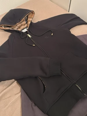 Bu*berry Svart hoodie guldig dragkedja - Snygg svart hoodie från Bu*berry med dragkedja och klassiskt rutigt foder i huvan. Har dragsko med metalländar, fickor fram och diskret broderad logga på bröstet. Perfekt för dig som gillar exklusiv streetwear och vill ha något stilrent men ändå unikt.